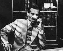 Black-and-white image of a man with a heavily bandaged nose sitting and talking on the phone. He wears a cream-colored suit and vest and boldly patterned tie; the collar of a white shirt is visible. Behind of him is a bookcase; in front of him, the edge of desk. A series of diagonal shadows descending from upper left falls over most of the image.