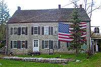 A large stone house with a large American flag draped across its upper right story behind an evergreen tree. In front of it is a blue and gfold historical marker.