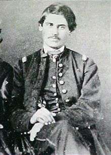 A white man with dark hair and a thin mustache sitting on a chair, his hands resting on his leg. He is wearing a military jacket and overcoat.