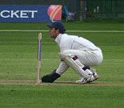 A wicketkeeper, side on, kneeling very closely behind the stumps, awaiting a delivery.