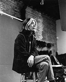 James Iha—a Japanese adult male with shoulder-length hair with white highlights—sits crossed-legged on a stool wearing a flannel shirt and jeans.