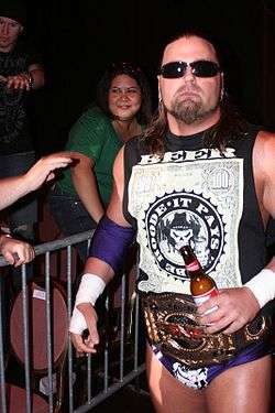 An adult white male wearing various colors of wrestling gear and a black white T-shirt as well as black sunglasses holding a beer bottle.