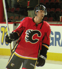Hockey player in red uniform, with a "C" in the middle. He leans to his left, holding his stick by his side.