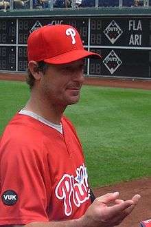 A man in a red baseball jersey with "Phillies" and red baseball cap with a white "P" on the front reaches his hand toward an unseen person.