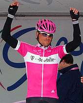 A man in a pink helmet wearing a pink top with black arm-warmers and gloves. He has hands above his head