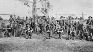 Black and white photo of Australian troops, armed with rifles, posing in a relaxed manner in front of a group of Japanese prisoners.
