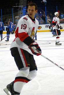 Hockey player in white uniform and helmet off. He skates across the ice, looking over his shoulder.