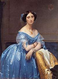 Princess Albert de Broglie wears a blue silk evening gown with delicate lace and ribbon trim. Her hair is covered with a sheer frill trimmed with matching blue ribbon knots. She wears a necklace, tasseled earrings and bracelets on each wrist.