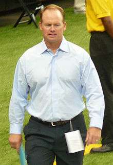 Candid photograph of Ireland walking on a football sideline wearing grey slacks and a light blue button-up shirt and holding a piece of paper in his right hand