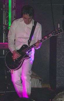 Jeff Schroeder—a Korean adult male—plays guitar in a white jumpsuit