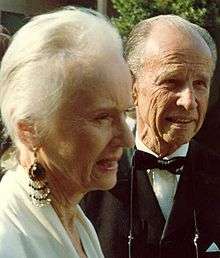 A photograph of an elderly woman seen from the side standing next an elderly man wearing a tuxedo. She is wearing a white dress and an earring.