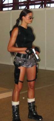 A full body picture of a dark-haired woman dressed in a black, sleeveless shirt and brown, camouflage shorts holding a silver gun.