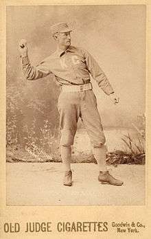 A baseball player is standing in his uniform, with his arm extended in the act of throwing a baseball.
