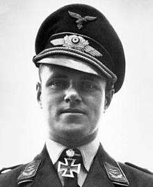 The head and shoulders of a young man. He wears a peaked cap and a military uniform with an Iron Cross displayed at the front of his shirt collar. His facial expression is a determined; his eyes are looking into the camera.