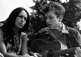 Outdoors,Joan Baez is sitting next to Bob Dylan who is playing an acoustic guitar, ca 1960s.