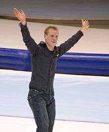 Jochem Uytdehaage on the ice, with his hands raised, he is dressed in street, not competition, clothes.