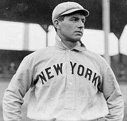 A waist-up photo of a man in a white baseball uniform reading "NEW YORK" across the chest and a white baseball cap. The man is looking to the right of the image.