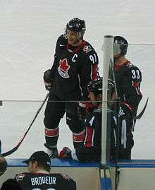 Hockey player in black uniform with a red leaf in the middle. He smiles and stands near the sideline of the rink.