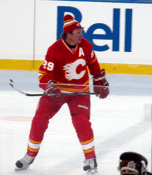 A hockey player in a red uniform with white and yellow trim and a stylized "C" logo on his chest skates forward as he looks to his left.