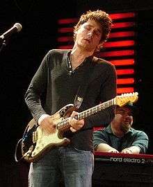 A Caucasian male wearing a long sleeved green shirt with jeans. The male is strumming on a guitar while holding the instrument with his two hands.