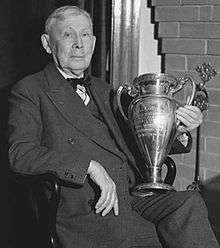 Elderly man, seated, wearing a suit and bow tie and holding a large trophy