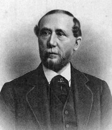 A black-and-white image of a man with thinning hair, mustache and beard wearing a suit and vest, shown from the chest up