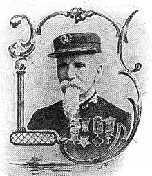 Framed portrait of a white man with a mustache and long beard wearing a flat-topped cap and a military jacket with four large medals hanging from ribbons on the left breast.