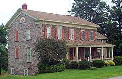A two story stone house with a beady-looking surface. It has reddish-orange wooden shutters and pale yellow trim. There is a lawn and several bushes in front.