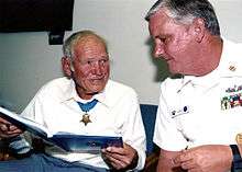 an older man wearing a medal of honor looks at a book while talking to another man wearing master chief summer whites