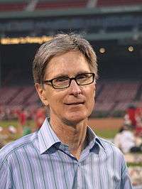 A portrait shot of a merry looking middle-aged Caucasian male (John W. Henry) looking straight ahead. He has short greying hair, and is wearing a blue-striped shirt with the top button open. In the background is a baseball stadium.