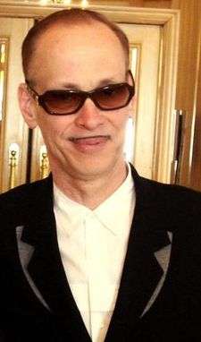 A balding man with a small mustache, in sunglasses and wearing a dark suit.