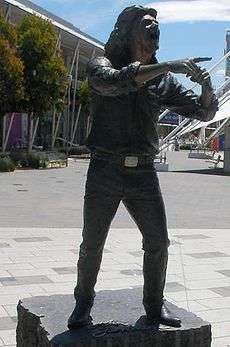 Statue of Farnham standing with a microphone in left hand at his opened mouth and pointing with right forefinger. Statue is on a block of stone with cursive lettering, John Farnham, in front of feet. Background includes a tiled area, wide footpath, trees and buildings.