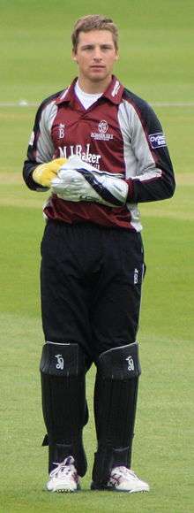 A young man with light brown hair is standing on some grass. He is wearing a burgundy, grey and black cricket uniform, large gloves and black trousers covered by black think pads on his lower legs.