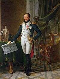 Painting shows a slightly balding man with dark hair standing in a room holding a building plan. He wears an early 1800s French military uniform: a blue coat with a white waist coat, white breeches, and black boots which rise above his knees.