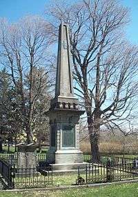 A stone obelisk and grave marker inside a low black fence in the middle of a cemetery. The obelisk is on an ornate square pedestal with a green metal plaque on it.