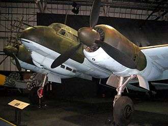 A color photograph of the front section of a twin engine propeller aircraft in a museum, shown in semi profile, viewed from the front left. The aircraft is painted green and light blue. Antennas are protruding from the nose of the aircraft.