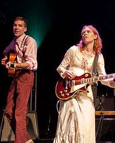 A skinny man in a white shirt and burnt red pants on stage stares into the camera while Welch next to him in a white dress focuses on playing her guitar. The man looks to be in his 20s.