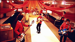 A seven-member band are playing instruments against the backdrop of orange, yellow, and red drapes.