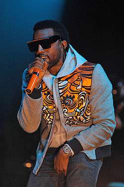 A man wearing sunglasses, blue jeans, a gray sweatshirt and an orange vest with tiger-stripes designs performs on a stage into an orange microphone.