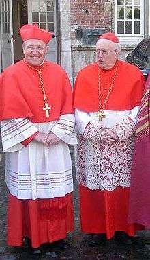 Cardinals Walter Kasper (left) and Godfried Danneels (right) wearing their choir dress: scarlet (red) cassock, white rochet trimmed with lace, scarlet mozetta, scarlet biretta (over the usual scarlet zucchetto), and pectoral cross on cord.