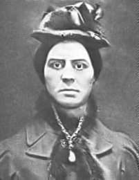 Head-and-shoulders view of a heavy-faced woman with dark hair, wearing a bonnet and a fur-lined coat, with a prominent necklace in front