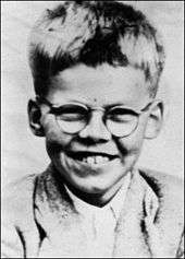 Head and shoulders monochrome photograph of a smiling short-haired young boy wearing spectacles.