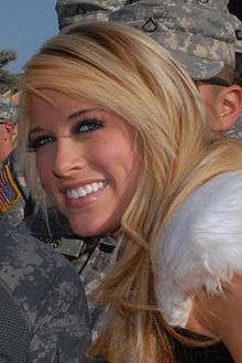 A close-up photo of a young, Caucasian blonde woman, who is smiling at the camera. Men in camouflage uniforms are visible in the background.