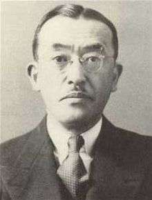 Close up of the head and shoulders of a solemn middle-aged Japanese man with a small mustache. He is wearing a suit, tie, and glasses.