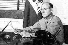 Man in shirt and tie sits at a desk. His shirt is neatly ironed. A telephone and writing pens are on the table. In the background is a US Army Corps of Engineers flag.
