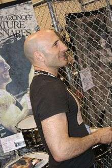 Kenny Aronoff—a middle-aged Caucasian male with a shaved head—smiles in a profile shot while standing in front of a drum set and posters of himself performing.
