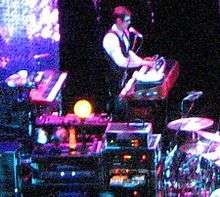Khristopher Pooley—a Caucasian male wearing a suit and sunglasses—plays keyboards onstage.