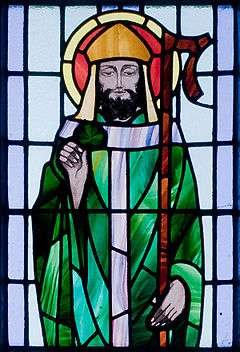 A stained glass window depicts Saint Patrick dressed in a green robe with a halo about his head, holding a sham rock in his right hand and a staff in his left.