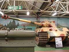 A frontal view of a large tank in a museum, painted pale yellow with some green and rust-brown blotches. Its curved-faced turret is turned to the left and the long gun overhangs the side by several meters.