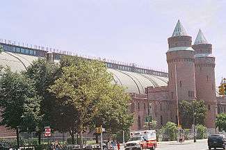 A large round-roofed building running the width of the image behind a city street and trees. At the right are two conical towers and an entrance pavilion